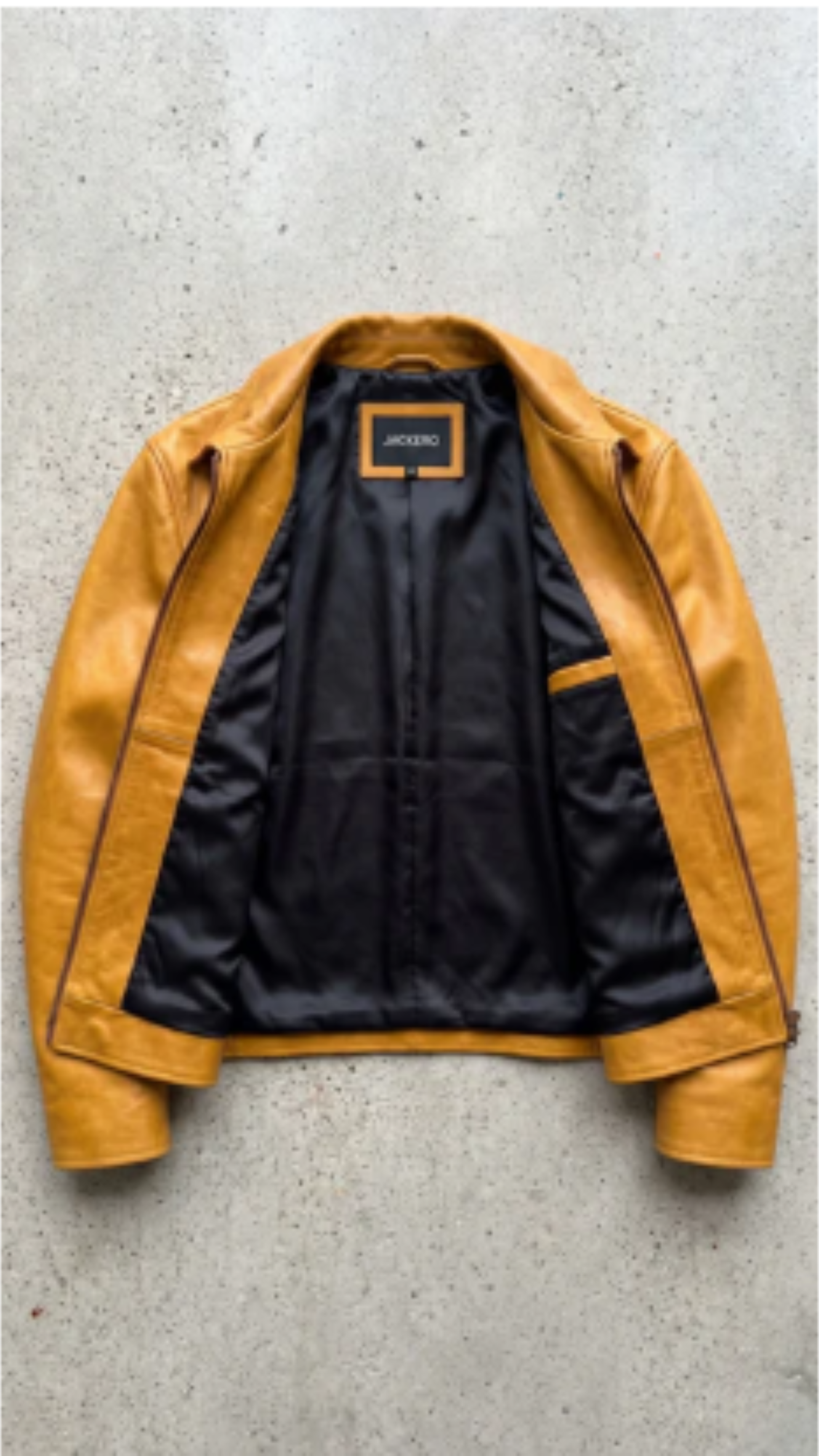 A mustard yellow leather jacket with a black satin lining, laid flat on a textured gray concrete surface. The brand label inside the collar reads 'JACKERO'.