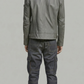 A man viewed from behind wearing a gray leather jacket, dark gray cargo pants, and brown boots, standing against a light gray studio background.