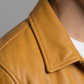 Close-up of a person wearing a mustard yellow leather jacket with a pointed collar over a white shirt, against a plain gray background.