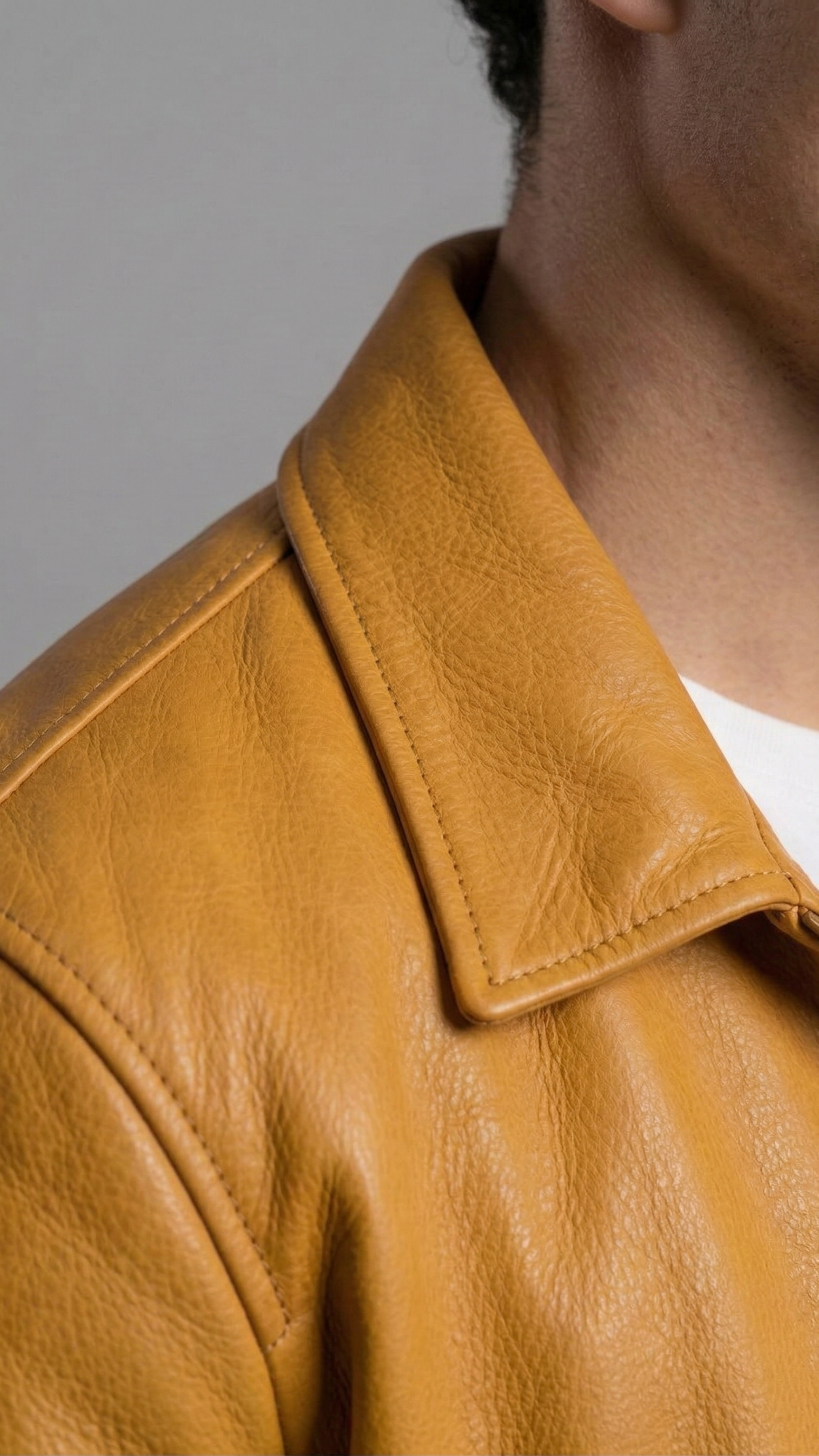 Close-up of a person wearing a mustard yellow leather jacket with a pointed collar over a white shirt, against a plain gray background.