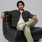 A man wearing a black leather jacket with cream stripes on the sleeves, blue jeans, and black boots, sitting on a wooden chair against a light gray background.