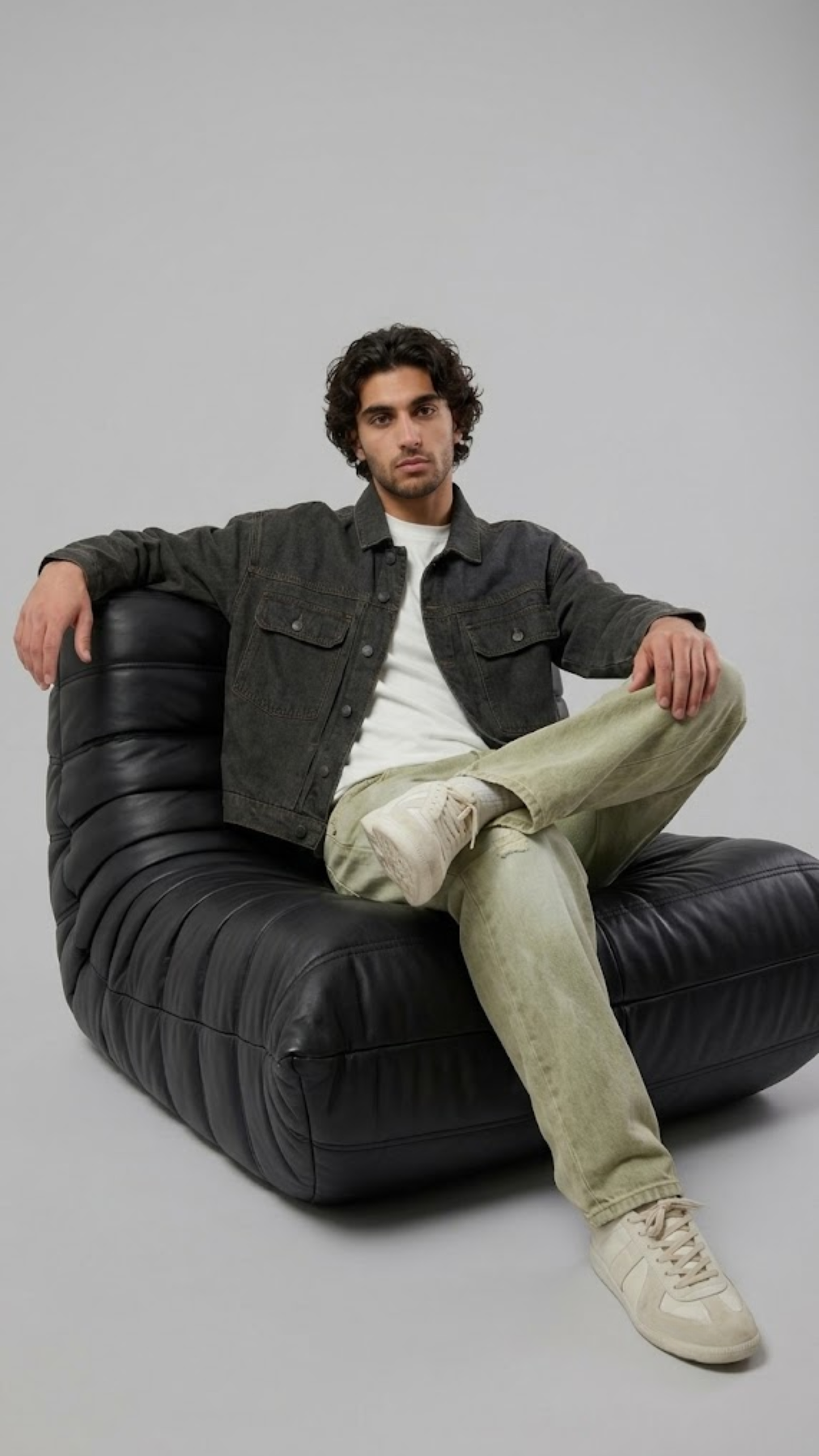A man wearing a black leather jacket with cream stripes on the sleeves, blue jeans, and black boots, sitting on a wooden chair against a light gray background.