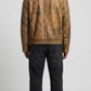 A man viewed from the back wearing a brown distressed leather jacket and dark charcoal jeans, standing against a light gray studio background.