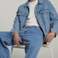 A young man sitting on a white chair wearing a light blue denim jacket, matching light blue jeans, a white t-shirt, and white sneakers against a light gray studio background.