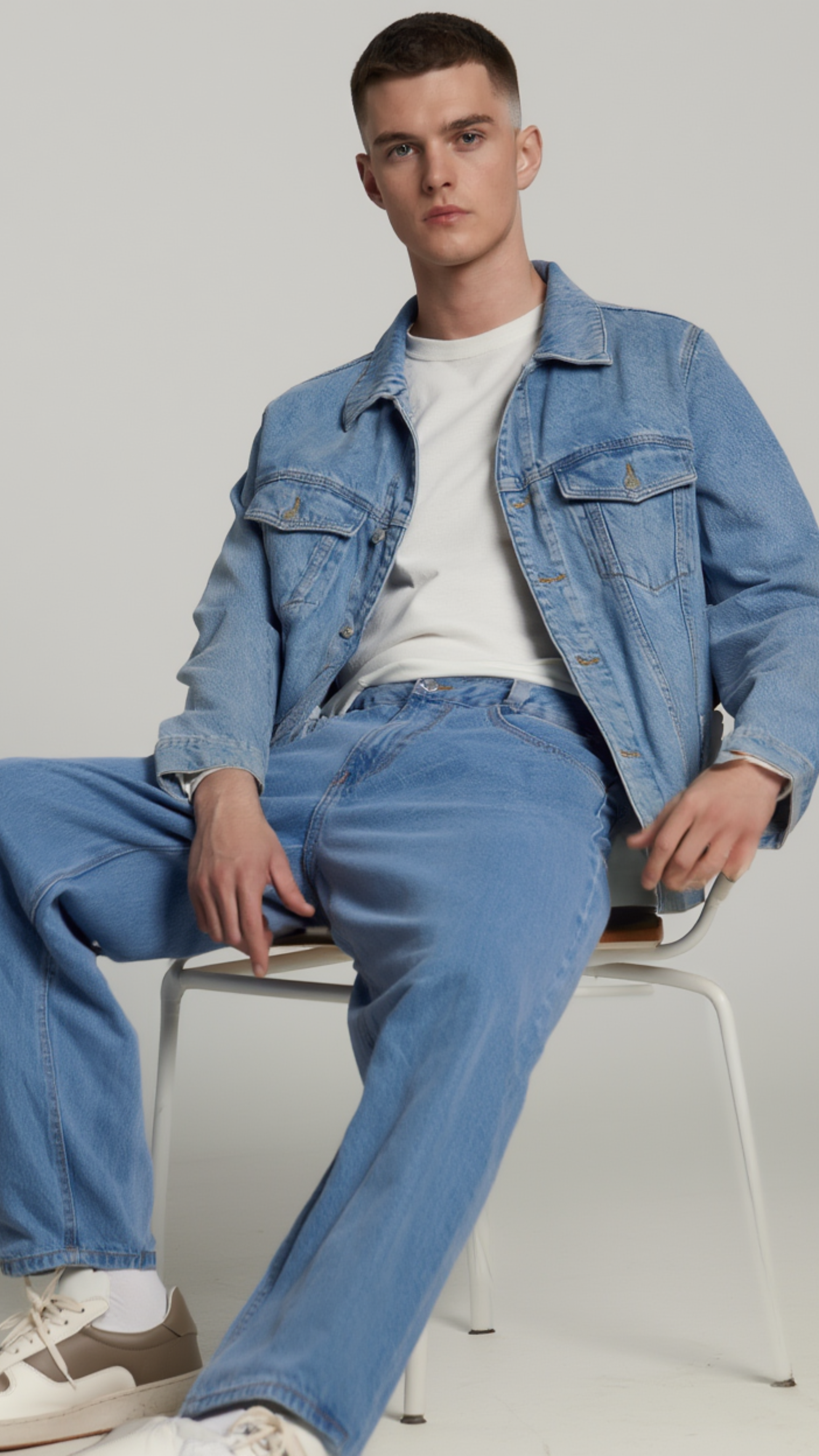 A young man sitting on a white chair wearing a light blue denim jacket, matching light blue jeans, a white t-shirt, and white sneakers against a light gray studio background.