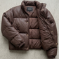 Close-up shot of a dark chocolate brown puffer jacket showing the premium textured fabric, zipper closure, and padded collar.