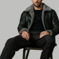 Man sitting on a chair wearing a black leather coat with gray fur lining against a plain background