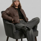Person sitting in a chair wearing a brown jacket, gray beanie, and gray pants on a plain background