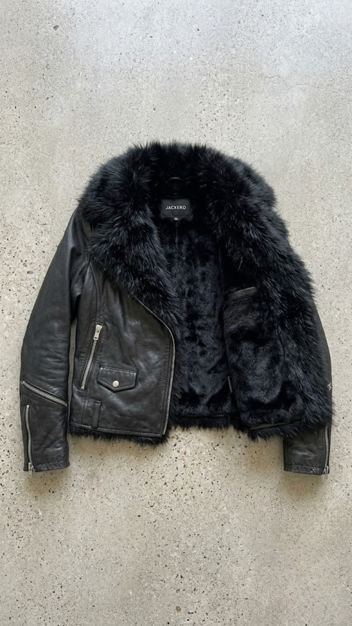 Studio flat lay photography of a high-quality black leather jacket with detachable black faux fur collar.