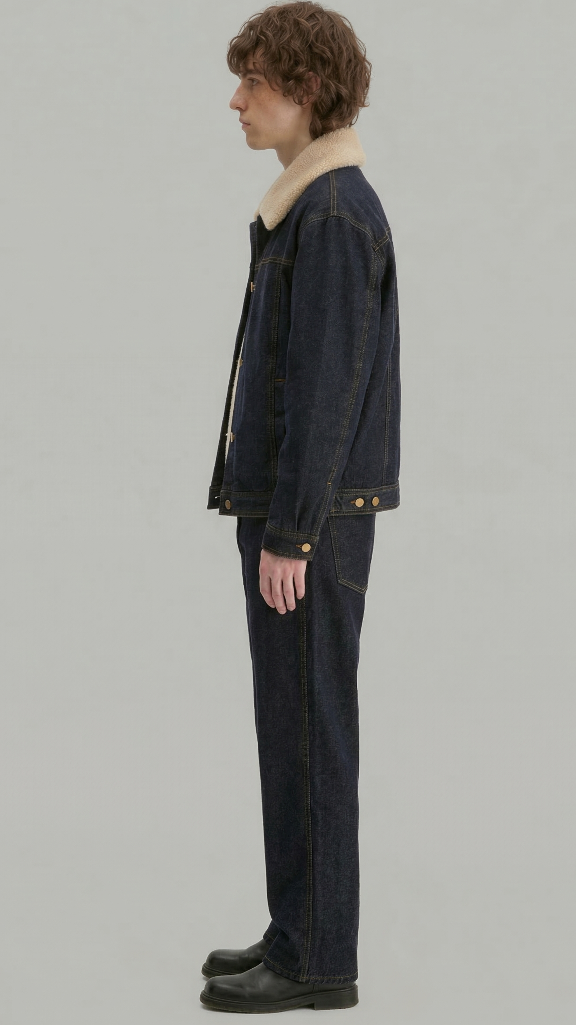 A model shown in profile wearing a dark indigo denim jacket with a beige shearling-style collar, matching dark denim jeans, and black leather shoes, against a solid light gray background.