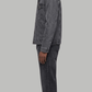 A man shown in profile wearing a dark charcoal gray washed denim jacket, light beige trousers, and beige sneakers, standing against a neutral light gray studio background.