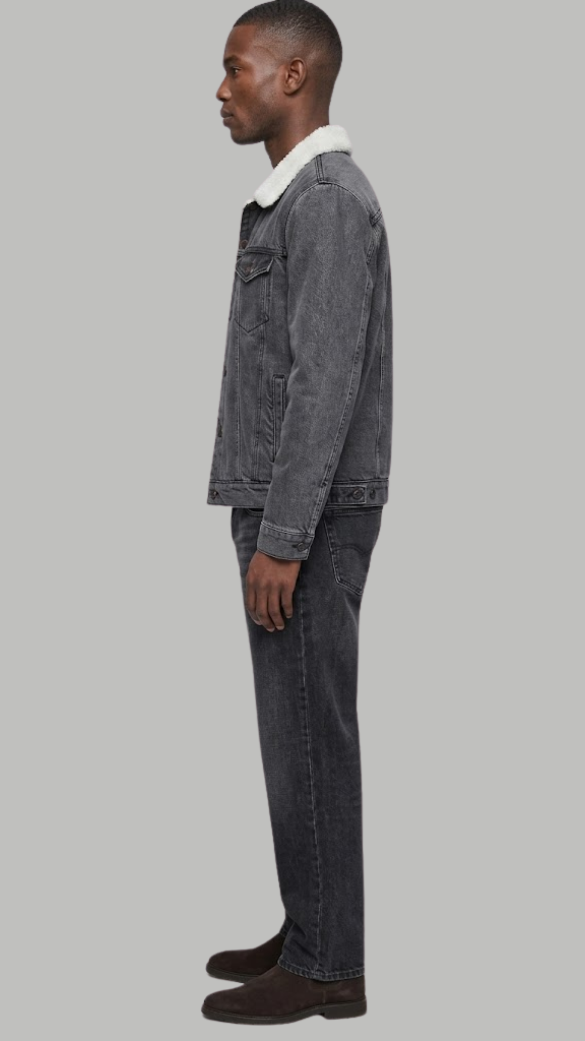 A man shown in profile wearing a dark charcoal gray washed denim jacket, light beige trousers, and beige sneakers, standing against a neutral light gray studio background.