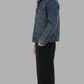 A man shown in profile wearing a washed dark gray denim jacket with a white collar, dark gray jeans, and brown shoes, standing against a solid gray background.