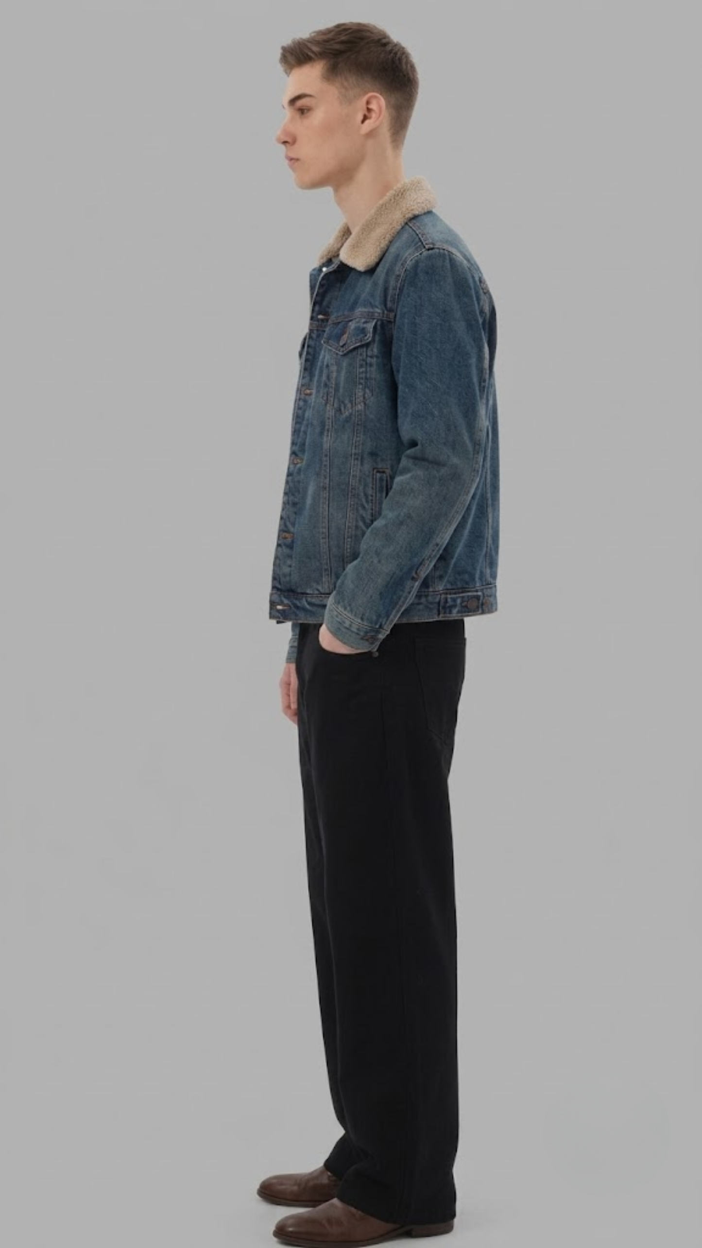 A man shown in profile wearing a washed dark gray denim jacket with a white collar, dark gray jeans, and brown shoes, standing against a solid gray background.