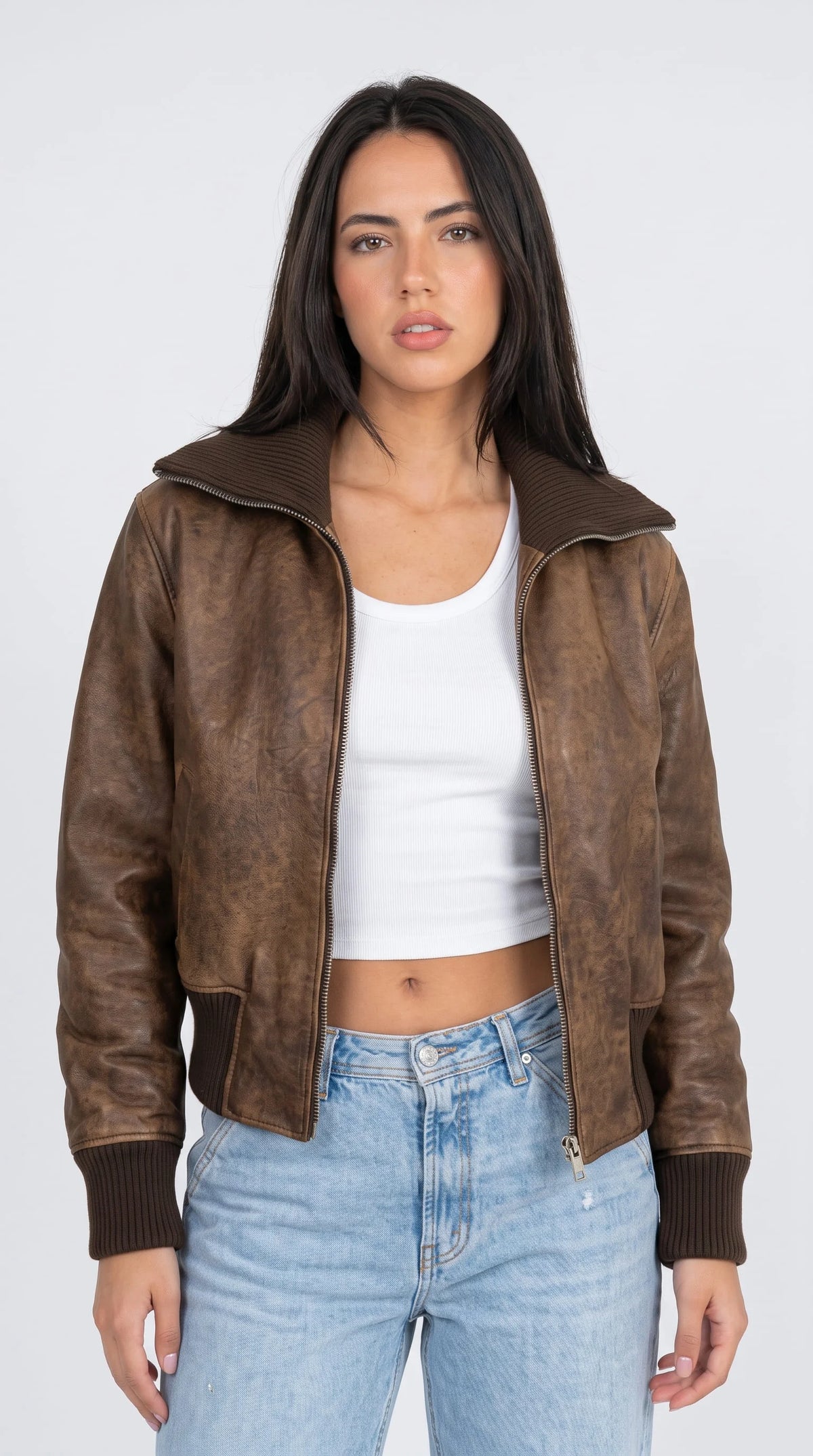 Women's distressed brown leather bomber jacket with ribbed knit collar and cuffs, styled with white crop top and light wash jeans