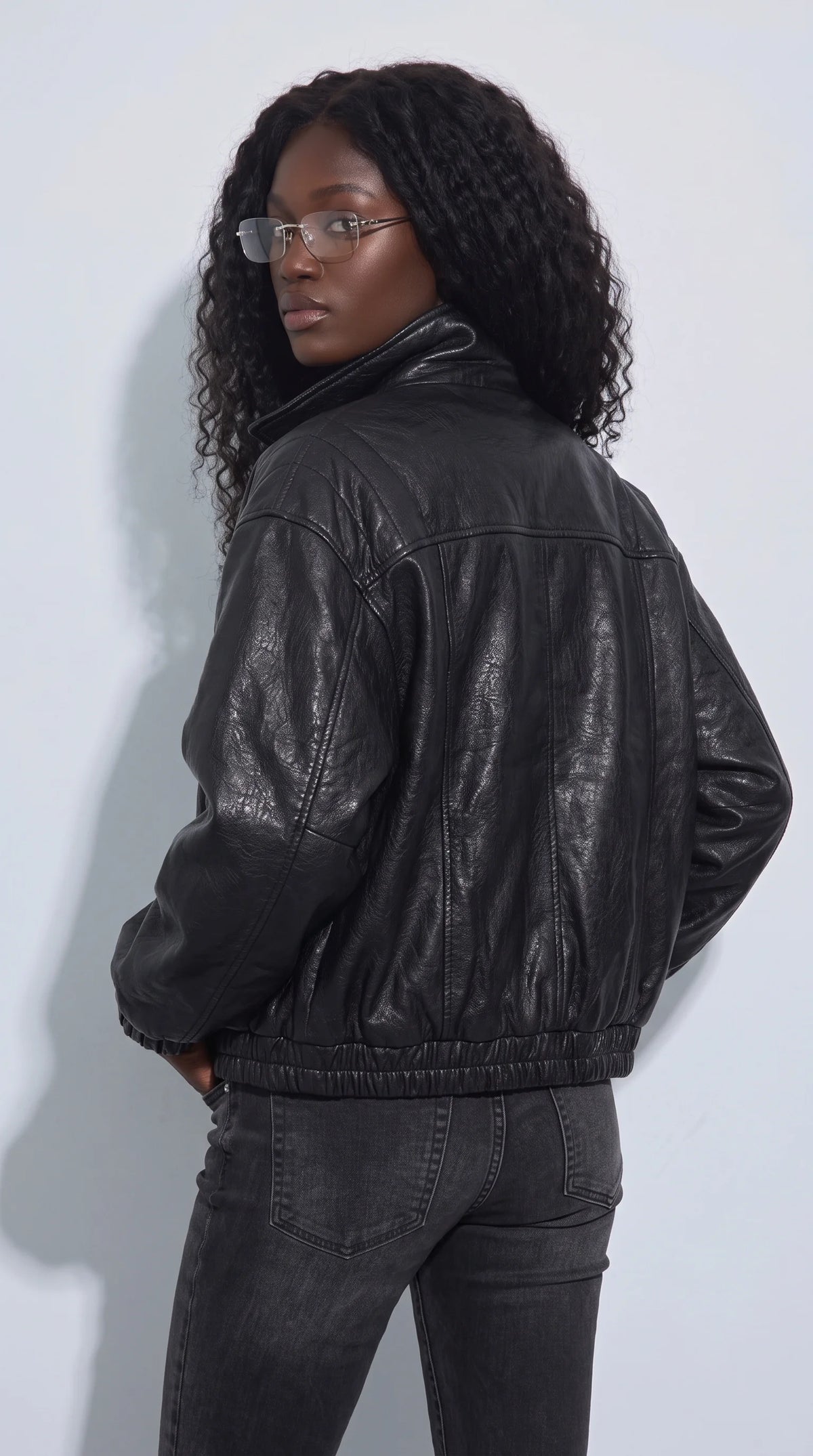 Women’s black cropped leather jacket with pronounced vertical panel stitching, curved shoulder seams and an elasticated waistband that shapes its relaxed bomber-inspired profile. The softly gathered hem and textured black leather create a strong silhouette over dark denim, offering a versatile statement layer for contemporary streetwear, casual weekend looks and minimalist wardrobes.