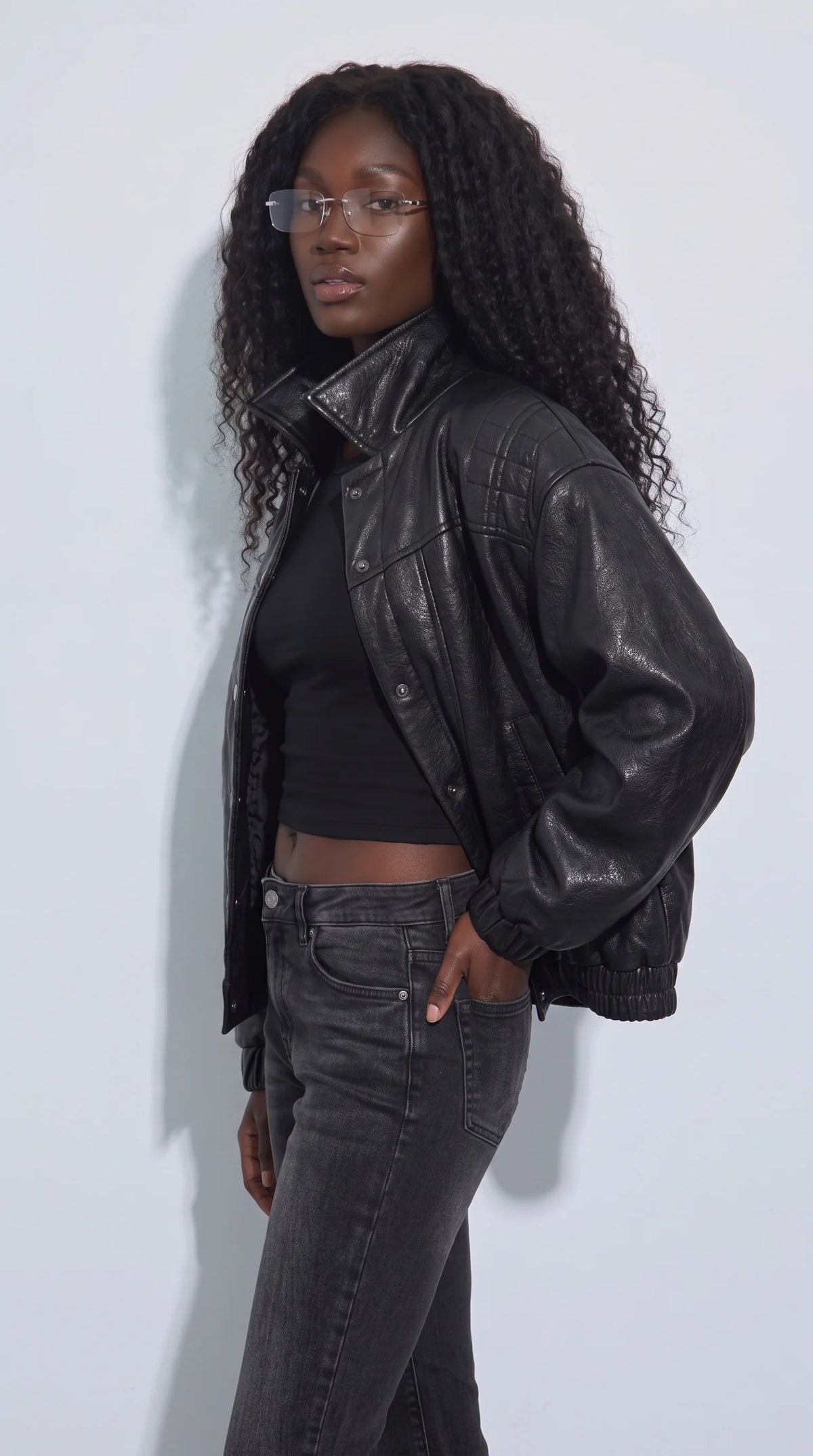 Black genuine leather short jacket worn open over a black crop top and charcoal denim, highlighting its wide stand-up collar, snap-button placket and softly voluminous sleeves. Quilted shoulder detailing adds a tailored accent to the relaxed cropped fit, creating a bold moto-inspired outfit for modern urban fashion, evening street style and layered monochrome looks.