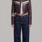 A model with curly dark hair stands against a light gray studio background wearing a cropped dark burgundy leather jacket with cream stripes on the chest and sleeves, dark blue jeans, a white top, and white sneakers.