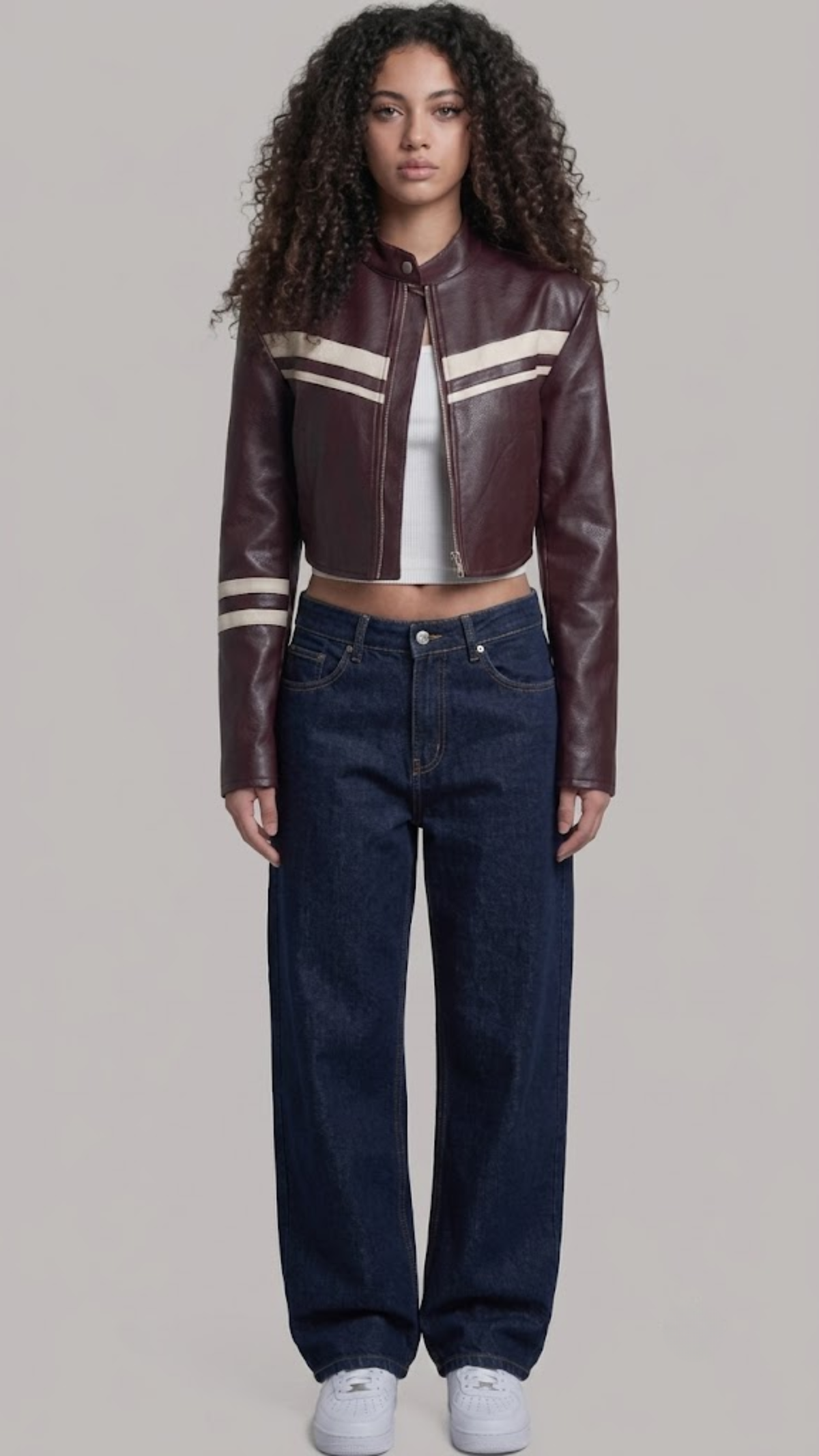 A model with curly dark hair stands against a light gray studio background wearing a cropped dark burgundy leather jacket with cream stripes on the chest and sleeves, dark blue jeans, a white top, and white sneakers.