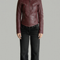 A model wearing a cropped, dark burgundy leather moto jacket with black jeans and white sneakers, standing against a solid gray studio background.