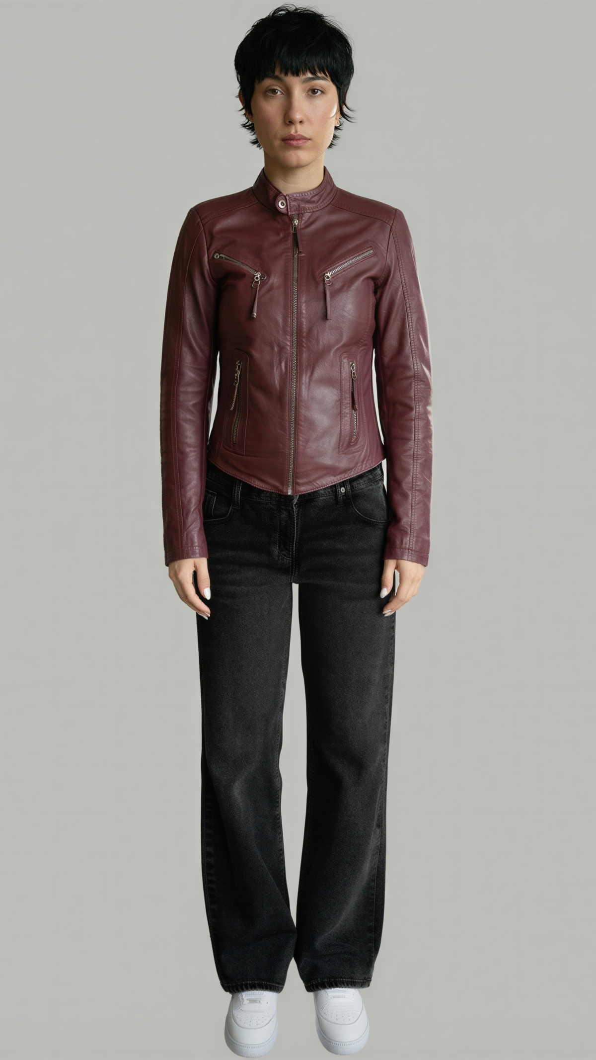 A model wearing a cropped, dark burgundy leather moto jacket with black jeans and white sneakers, standing against a solid gray studio background.