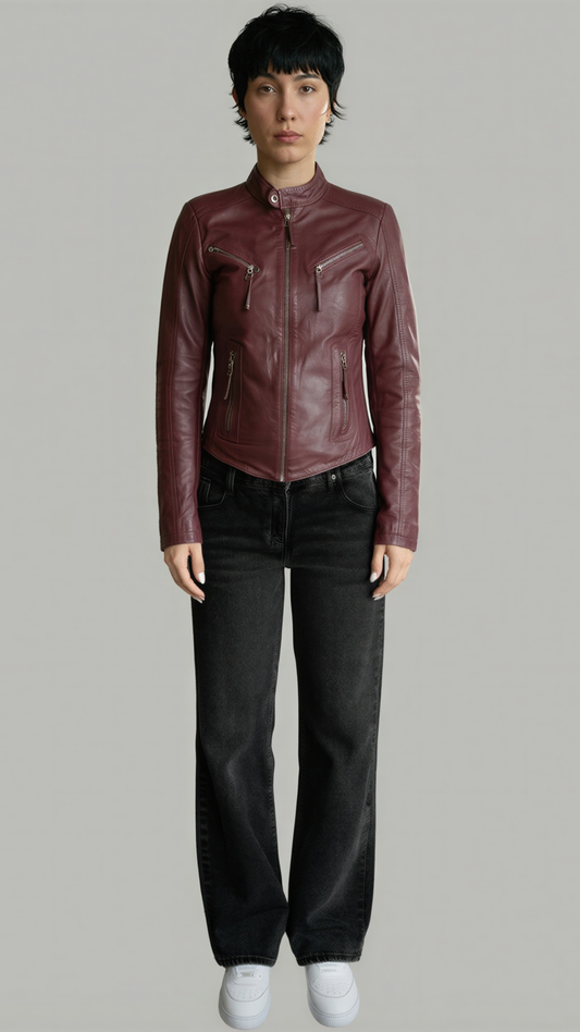 A model wearing a cropped, dark burgundy leather moto jacket with black jeans and white sneakers, standing against a solid gray studio background.