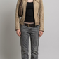 A model with curly blonde hair stands against a light gray studio background wearing a beige leather jacket, a black top, gray jeans, and brown shoes, with a brown shoulder bag.