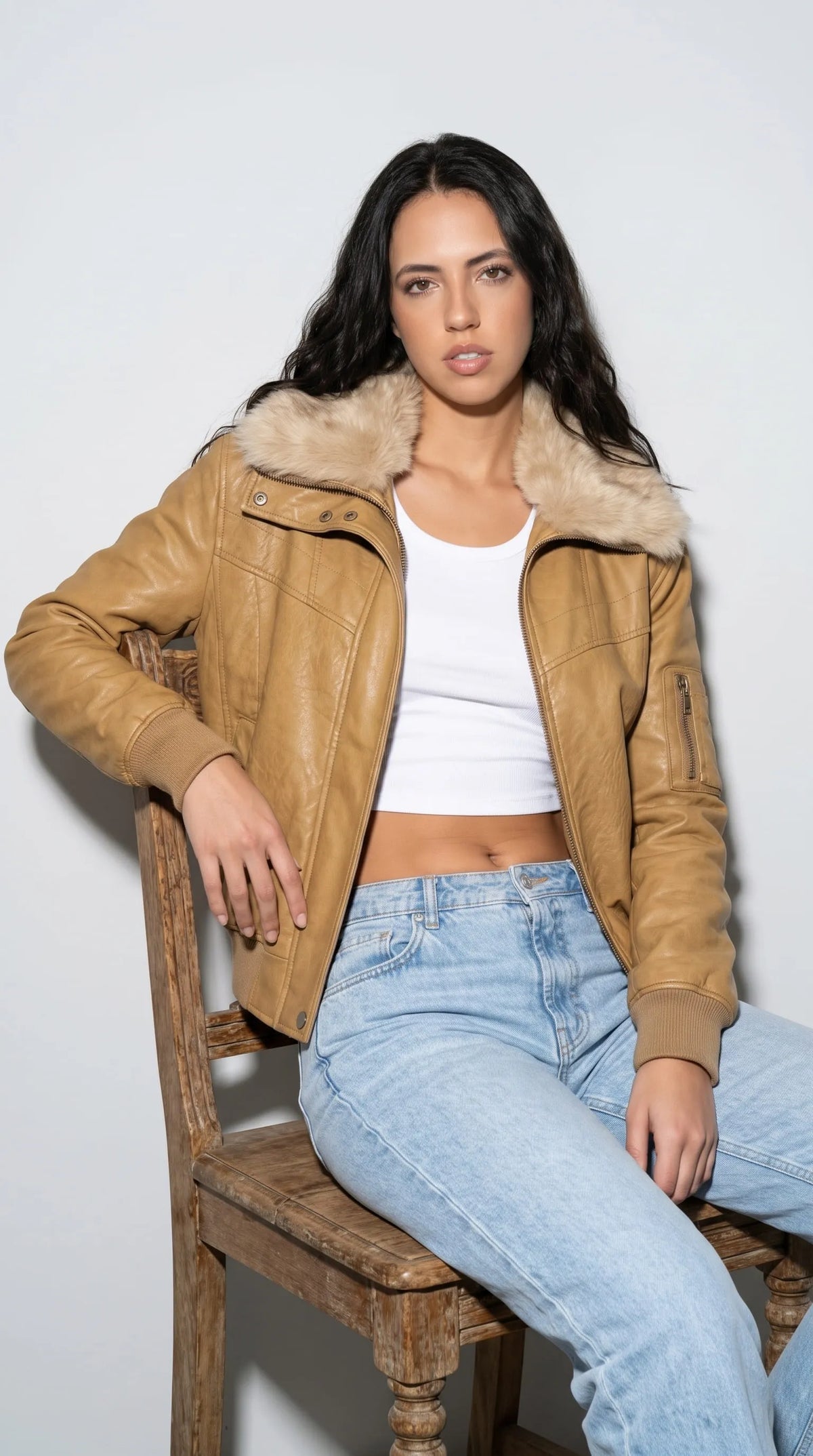 Relaxed fashion styling featuring a golden mustard leather bomber jacket with a fluffy cream collar, worn with a white crop top and light-blue jeans while seated on a rustic wooden chair. The cropped cut, ribbed cuffs and warm leather finish create an easy vintage-inspired outfit that balances casual denim dressing with a bold statement layer for everyday fashion and weekend streetwear.