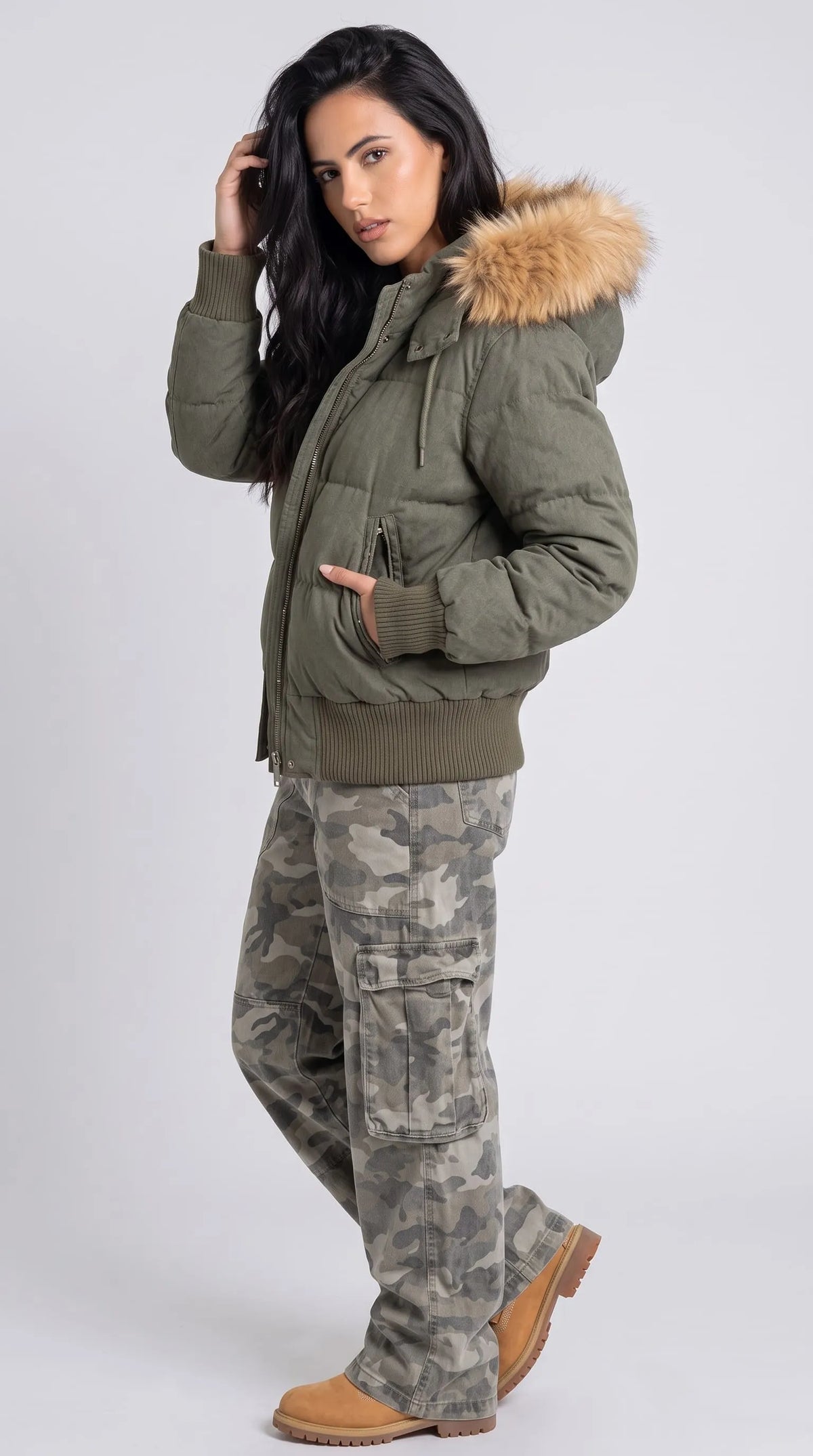 Women's oversized olive puffer jacket featuring a brown faux-fur-trimmed hood, padded sleeves, a zip-and-snap front closure and ribbed cuffs and hem, styled with loose camouflage cargo pants and tan lace-up boots. This balanced winter outfit pairs a voluminous quilted jacket with practical utility-inspired pieces, creating a distinctive casual look for outdoor outings, travel and urban streetwear.
