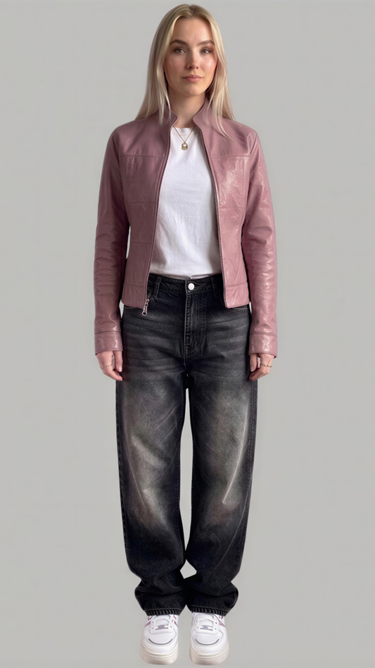 A woman modeling a dusty pink leather jacket over a white t-shirt and dark gray washed jeans, paired with white sneakers, against a solid gray background.